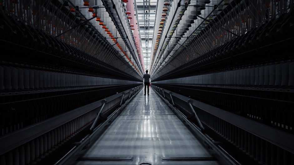 Long dark spinning hall with banks of bobbins converging on a single distant figure