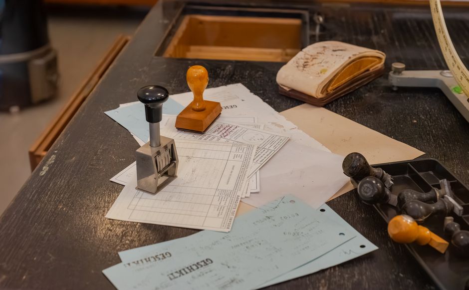 A records desk with a numbering stamp, a rubber stamp and old printed forms in a dark tray