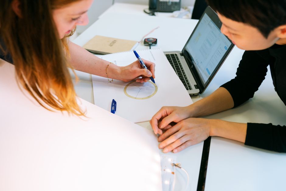 Two people at a desk, one sketching a diagram beside an open laptop